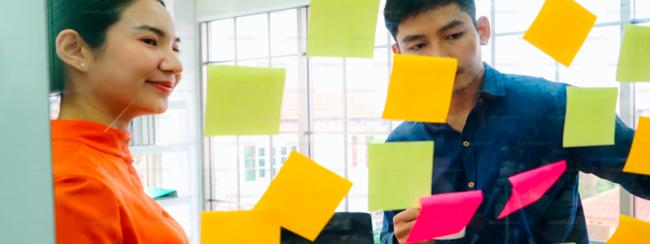 Two adults organizing Post-It notes on glass wall
