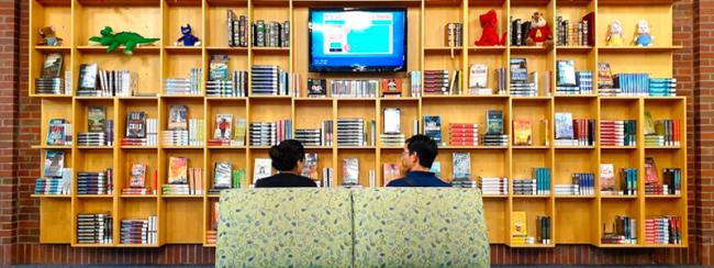 Two adults sit in chairs facing a huge array of resources, books and a wall mounted screen on open shelves.